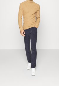 Beiger, strukturierter Rollkragenpullover, marineblaue Slim-Fit-Hose und weiße Sneaker. Das Outfit zeichnet sich durch ein geripptes Strickmuster und glatten Stoff aus.