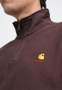 Polaire bordeaux à fermeture éclair partielle avec un emblème jaune et un logo brodé rouge. Texture lisse avec des coutures plates et un col montant.