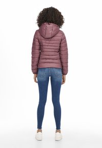Kurze Pufferjacke in Mauve, mit gestepptem Design, Kapuze und elastischen Bündchen, kombiniert mit dunkelblauen Jeans und weißen Sneakers.