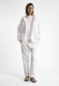 Haut à capuche blanc avec fermeture éclair et poches, porté par-dessus un t-shirt blanc, associé à un pantalon de survêtement blanc. Design minimaliste et texture douce. Chaussures brunes à enfiler.