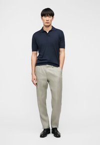 Hombre con polo azul marino de manga corta, pantalones beige plisados y zapatos de vestir negros de pie contra un fondo blanco liso.