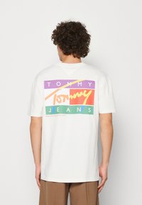Camiseta de algodón blanca con un gráfico colorido en la parte trasera que presenta las palabras "TOMMY JEANS" en varios colores vivos y formas geométricas.