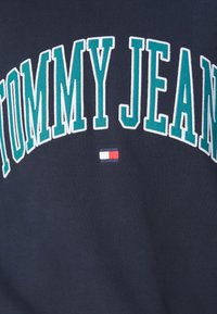 Sudadera azul marino con "TOMMY JEANS" en grandes letras bordadas en verde, con un pequeño detalle del logotipo en rojo, blanco y azul en el centro.