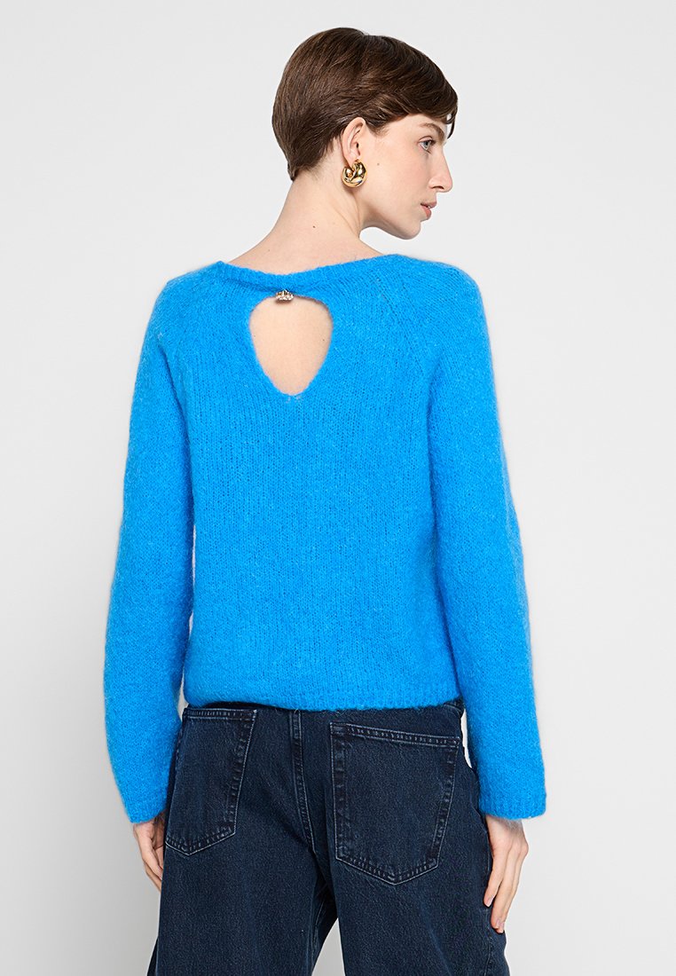 Donna di profilo che indossa un maglione lungo a maniche nel brillante colore blu, con un'apertura a chiave e una chiusura decorativa sulla parte alta della schiena, abbinato a jeans scuri.