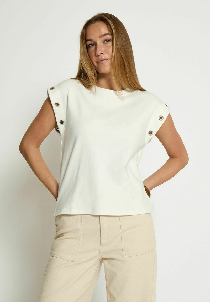 Top bianco a maniche corte con spalle arrotondate, presenta dettagli in metallo sui lati delle maniche, realizzato in un tessuto morbido, abbinato a pantaloni beige.