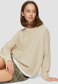 Beige, gerippter Strickpullover mit Rundhalsausschnitt und überschnittener Passform. Verfügt über abfallende Schultern und strukturierten Stoff, getragen über grünen Karoshorts.
