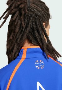 Jersey atlético azul con acentos naranjas, que presenta un logo redondo en la parte posterior y un cuello alto. La tela parece texturizada y transpirable.