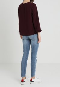 Frau, die mit dem Rücken zur Kamera steht, trägt eine dunkelburgunderfarbene Bluse mit langen Ärmeln und Rüschensaum, hellblaue Jeans und weiße Sneaker mit roten Details.