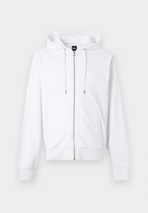 Mand står op ad hvid væg iført hvid hættetrøje med lynlås, sort skjorte, beige ternede løse bukser og sorte sko med hænder i lommerne.