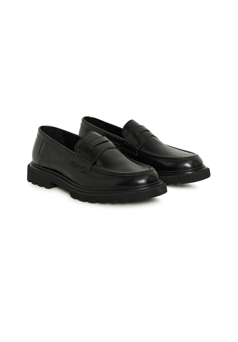 Paire de mocassins noirs en cuir à enfiler avec semelles épaisses à rainures et bride décorative sur le dessus, présentée sur fond blanc.