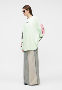 Frau mit Sonnenbrille, übergroßem, hellgrünem Langarmhemd mit Text auf Ärmeln und Brust sowie einem Patchwork-Denim-Maxirock.