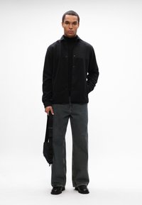 Casaco de lã preto com botões e colarinho, usado sobre uma camisola preta de gola alta; combinado com umas calças de ganga cinzentas de perna larga e sapatos pretos.