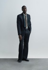 Melns uzvalks ar satīna atloka apkakli, kombinēts ar rūtainu kreklu un rakstainu kaklasaiti. Apavi ir spīdīgas melnas ādas. Vienveidīga pelēka fona.