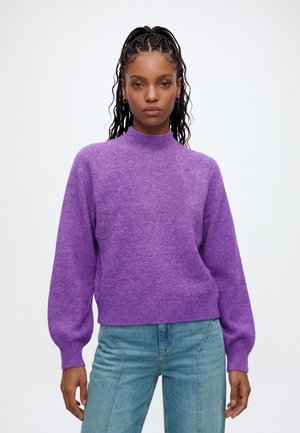 Kvinde med fletninger iført en lilla langærmet sweater og lyseblå jeans, stående foran en hvid baggrund.