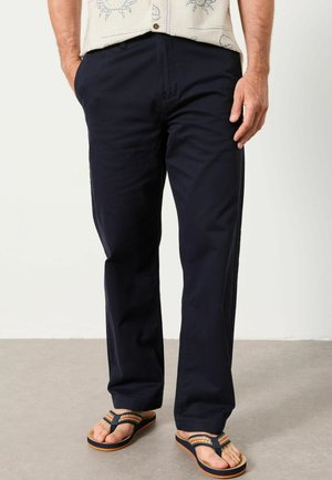 Mann trägt dunkelblaue gerade geschnittene Hose, ein cremefarbenes Hemd mit Sonnenmuster und gestreifte Flip-Flops und steht auf einem grauen Boden.