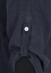 Marineblaue Leinenbluse mit aufgerolltem Ärmel, ausgestattet mit einem weißen Knopf und Nahtdetails. Der Stoff hat ein strukturiertes, atmungsaktives Aussehen.