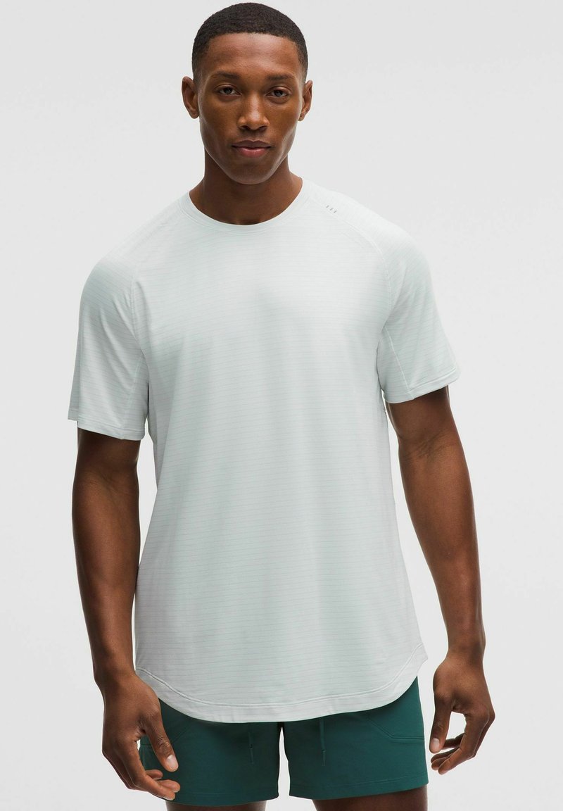 Muž s vypracovanou postavou, oblečený v svetlosivej športovej tričku s krátkym rukávom a tmavozelených šortkách, stojaci pred jednofarebným sivým pozadím.