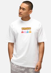 Camiseta blanca de manga corta con un gráfico colorido de "PAC-MAN" que presenta a cuatro fantasmas en colores rosa, azul y naranja. Diseño simple y casual.