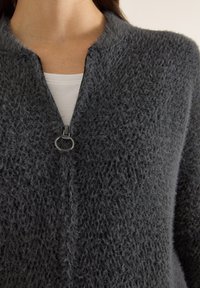 Šedý, chlupatý cardigan s vysokým límcem a zapínáním na zip. Má kulatý stříbrný přívěsek na zipu. Nosit přes bílou košili.