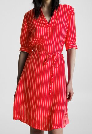 Czerwona sukienka do kolan z białymi pionowymi paskami, z rękawami do łokcia, dekoltem w serek i przewiązanym paskiem w talii, noszona przez osobę o ciemnych włosach.