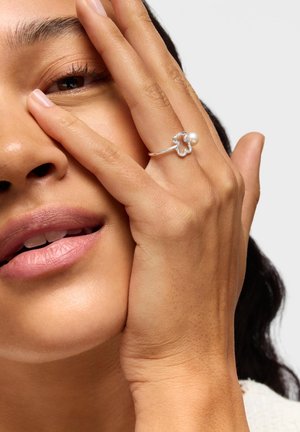 Primo piano del volto di una donna con la mano che copre parzialmente un occhio, indossa un anello d'argento a forma di orso con una piccola perla.