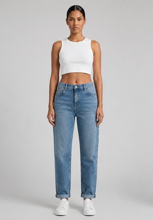 Kobieta stojąca przodem, ubrana w biały bezrękawnik typu crop top, wysokie niebieskie dżinsy z mankietami i białe trampki na szarym tle.