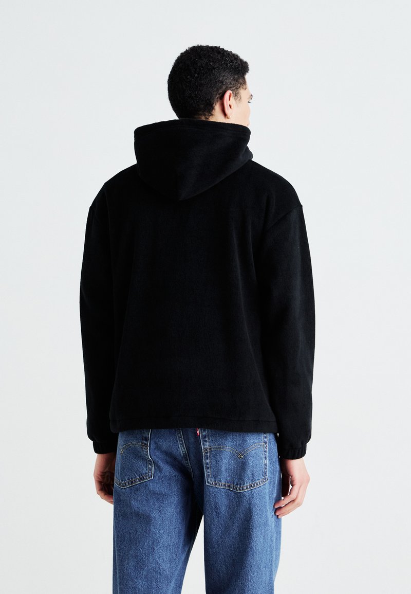 Haut de hoodie en polaire noir avec une grande capuche, coupe ample et poignets côtelés, associé à un jean en denim bleu clair avec des poches arrière.