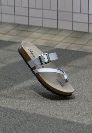 Sandale albă cu talpă de plută, curea cu cataramă metalică și buclă pentru deget, odihnindu-se pe asfalt lângă un perete acoperit cu plăci.