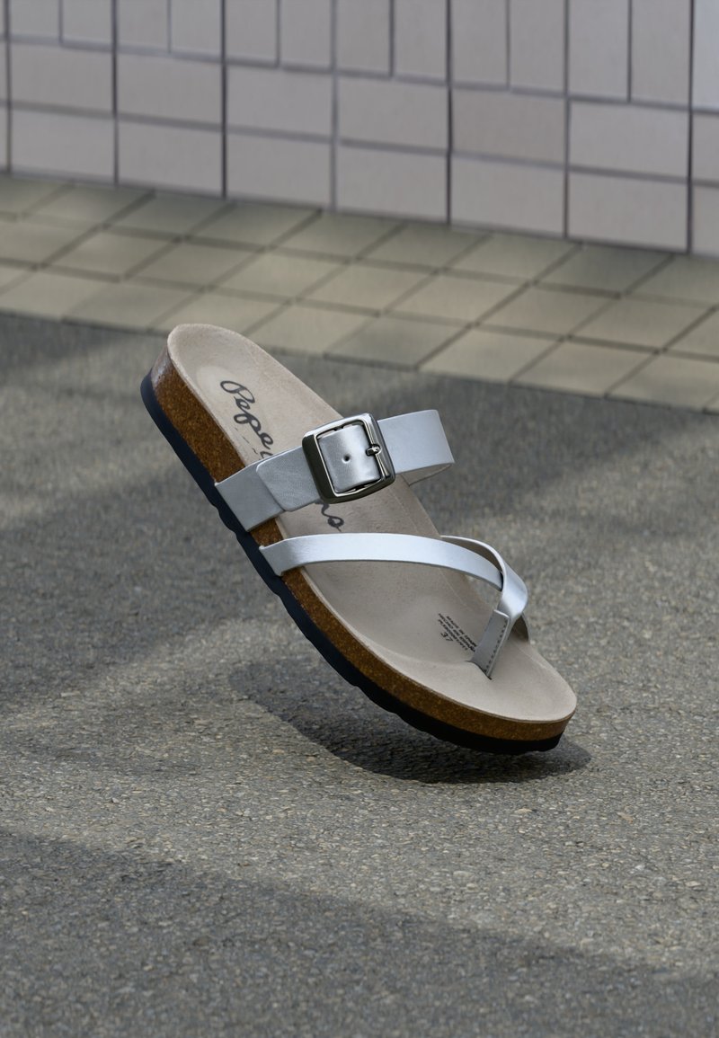 Λευκό σανδάλι με φελλό πάτο, μεταλλικό λουράκι με αγκράφα και θηλιά για δάχτυλο, ακουμπισμένο σε άσφαλτο κοντά σε τοιχο με πλακάκια.