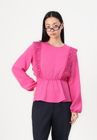 Haut rose à manches longues avec des accents en dentelle sur les épaules, taille froncée et tissu texturé ; associé à un pantalon sombre à jambes droites.