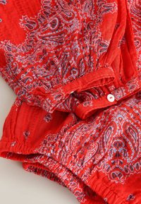 Tissu rouge avec un motif cachemire bleu et blanc, comprenant un bouton blanc et des détails de poignet élastiqué.