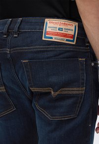 Σκούρο μπλε τζιν με κίτρινη ραφή και ορθογώνια ετικέτα με την επιγραφή Diesel Industry στη πίσω μέση.