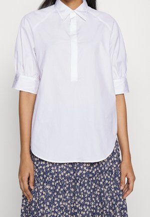 Femme portant un chemisier blanc à manches courtes avec col, associé à une jupe plissée bleue marine à motifs floraux.