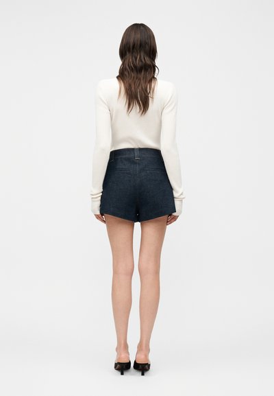 Γυναίκα με μακριά καστανά μαλλιά, φοράει λευκό μακρυμάνικο μπλουζάκι, σκούρα μπλε τζιν σορτς και μαύρα ψηλοτάκουνα παπούτσια, στέκεται με την πλάτη προς το μέρος μας.