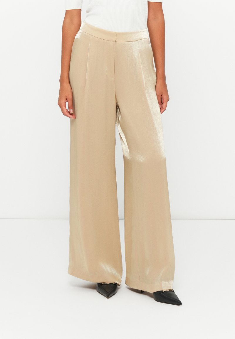 Weite Hosen aus einem metallisch goldenen Stoff mit glatter Textur. Mit einem taillierten Bund und scharfen vorderen Falten versehen. Kombiniert mit schwarzen spitzen Schuhen.