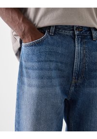 Modré džíny z denimu s vybledlým efektem, klasickým pětipokládkovým designem a kontrastním šitím, nošené s světlou košilí.