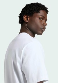 Maglietta bianca in cotone con scollo tondo e maniche corte, indossata da una persona con capelli neri corti e strutturati, su uno sfondo neutro.