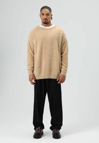 Beige, pluizige trui met brede halslijn, gecombineerd met losse zwarte broek en zwarte schoenen met rode veters. Minimalistisch ontwerp.