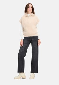 Beige nepbont vest met een ritssluiting aan de hals, gedragen over een wit langetermijn shirt, gecombineerd met donkere wijde jeans en crèmekleurige enkellaarsjes.