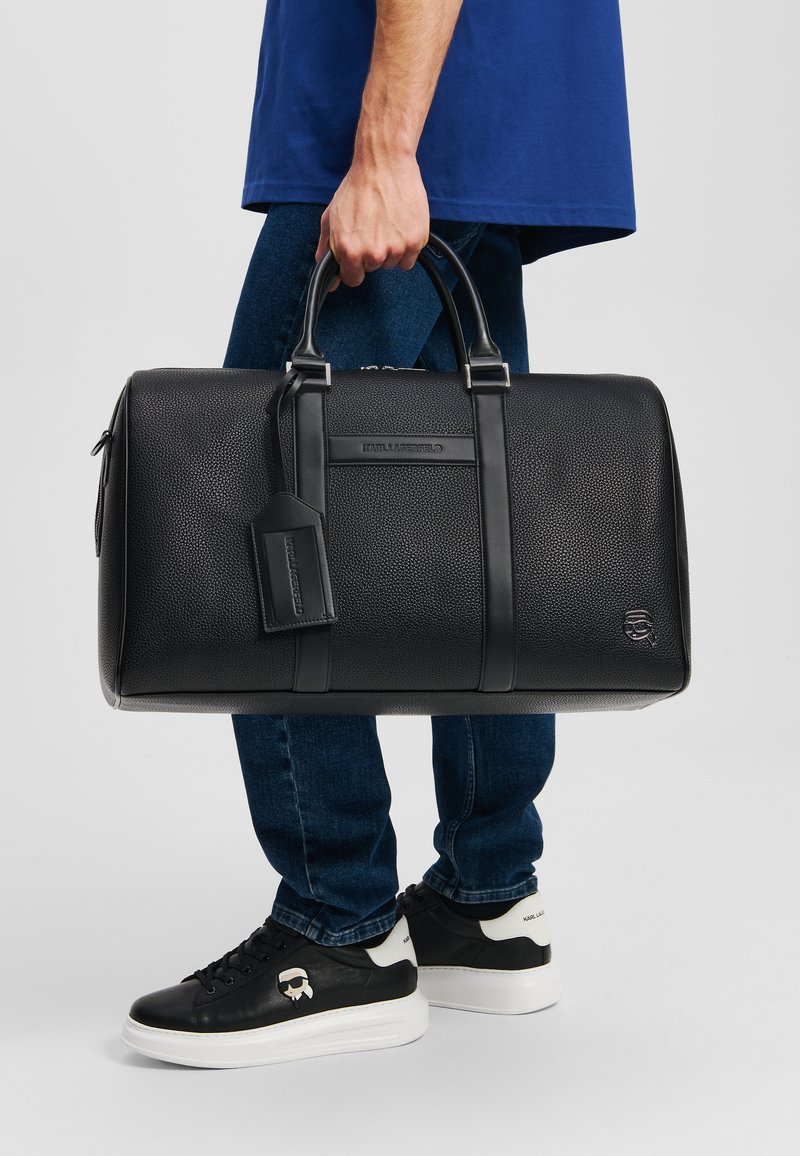 Sac de sport en cuir noir avec une surface texturée, deux poignées, une étiquette de bagage et un détail de marque. Accompagné de baskets noires avec des accents blancs.