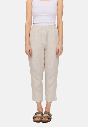 Woman wearing beige cropped linen pants with elastic waist, white sleeveless top, and olive green buckle sandals standing against white background.