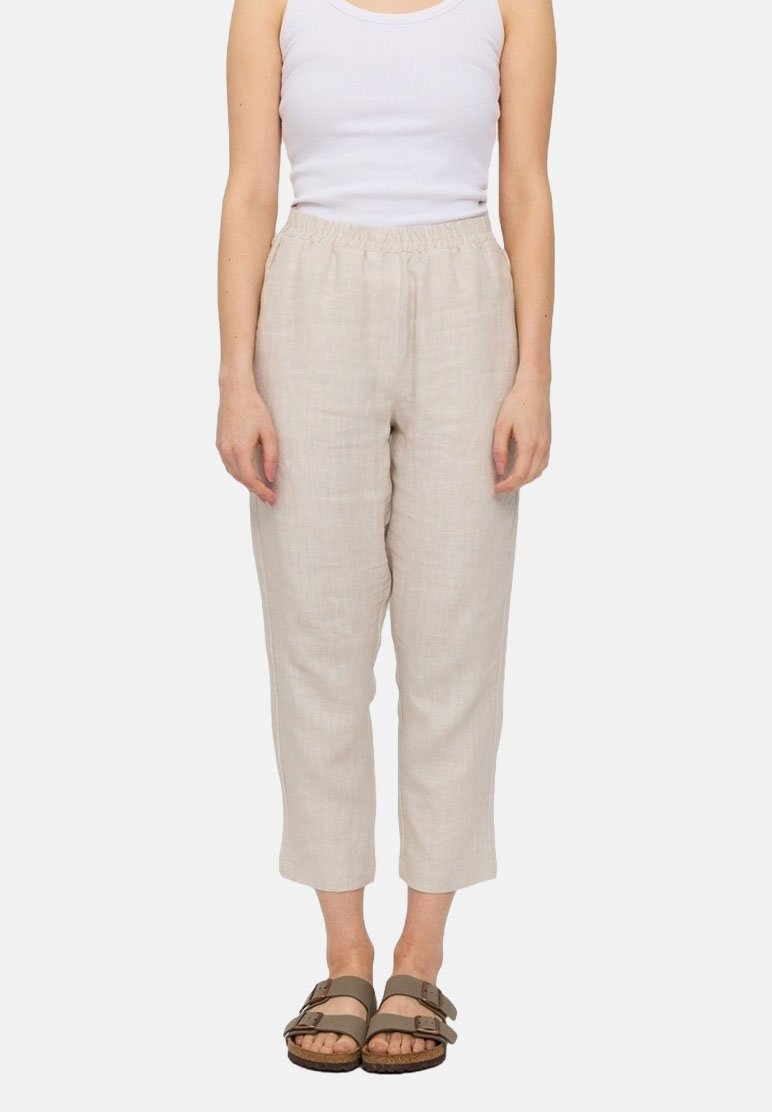 Woman wearing beige cropped linen pants with elastic waist, white sleeveless top, and olive green buckle sandals standing against white background.