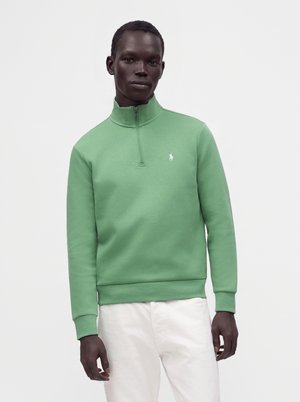Homme portant un sweat-shirt vert clair à fermeture éclair quart de zip avec un petit logo blanc sur la poitrine et un pantalon blanc, debout devant un fond uni.