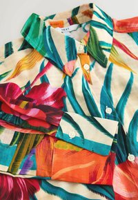 Chemise boutonnée avec un imprimé floral tropical vif en rouge, vert, orange et bleu sur fond crème.
