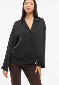 Svart silkebluse med lange ermer, knappelukking og delikat blondedetalj i kanten nederst. Kombinert med mørkebrune bukser.