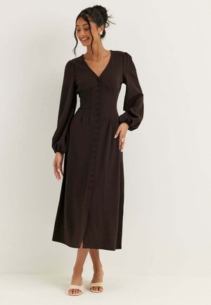 Donna che indossa un lungo vestito nero con bottoni, maniche a sbuffo e tacchi beige con punta aperta, in piedi davanti a uno sfondo bianco uniforme.