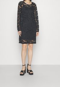Černé krajkové šaty s dlouhými rukávy, květinovým vzorem, kulatým výstřihem a rovnými lemy. Nosené s černými sandály na blokovém podpatku.