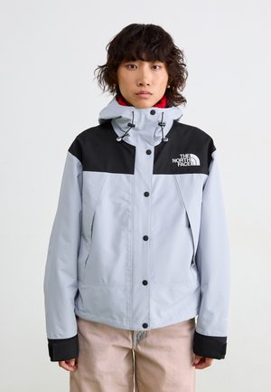 Persoon in een lichtgrijze en zwarte North Face-jack met capuchon, rode binnenlaag en lichtbeige broek, staande tegen een effen witte achtergrond.