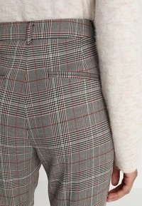 Pantalons à carreaux gris avec des motifs pied-de-poule, des accents rouges et une poche arrière. Le matériau semble texturé et tissé.