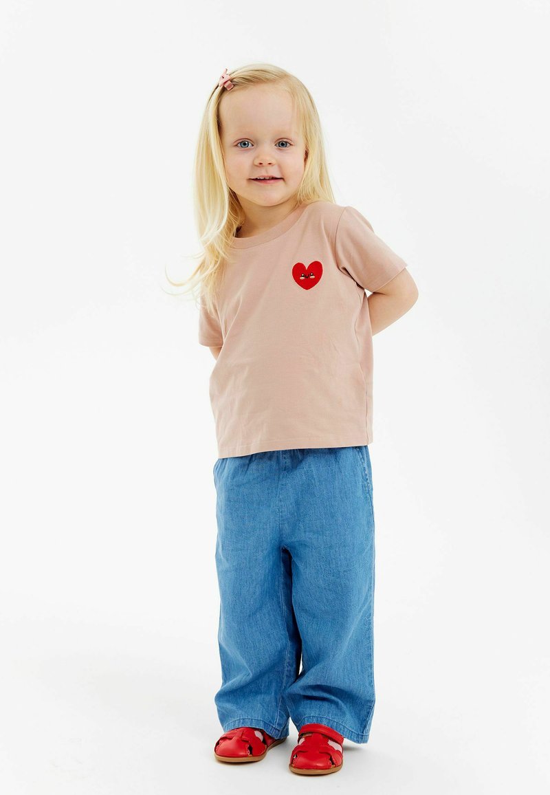 Ljusrosa t-shirt med kort ärm och röd hjärt-design, blå byxor med vida ben och röda sandaler. Långt blont hår stylet med en rosa klämma.